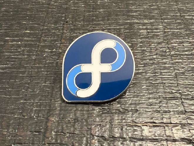 An enamel pin of the Fedora logo resting on a black wooden table.